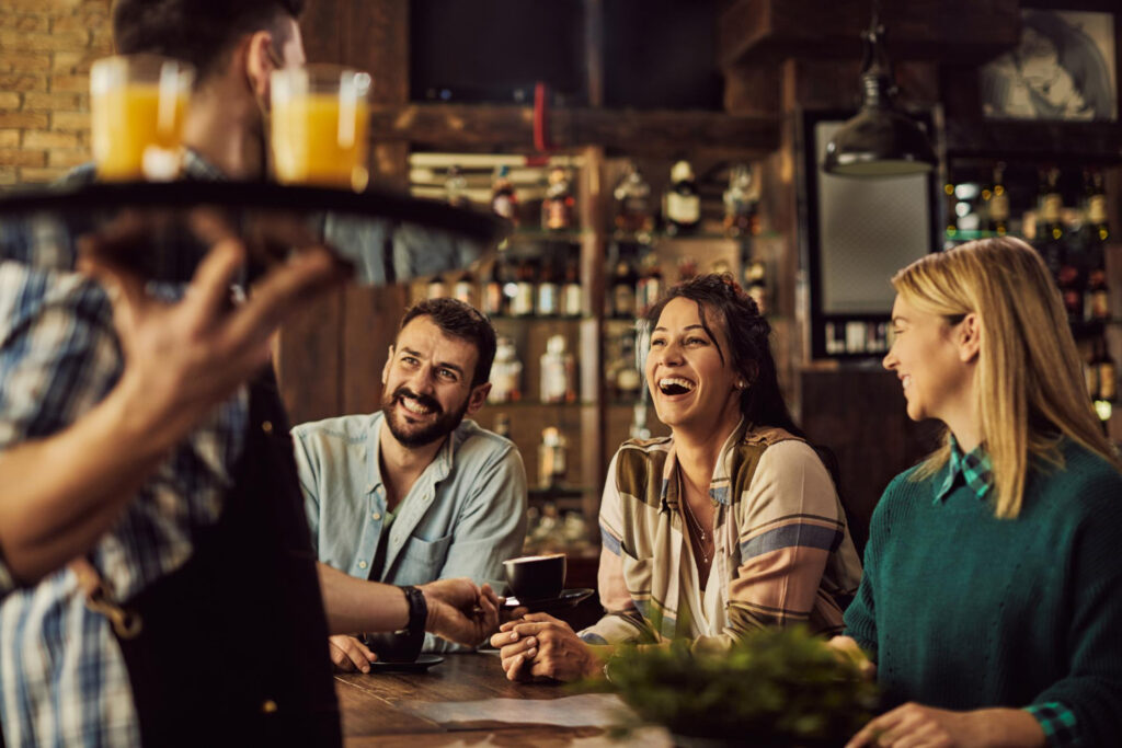 Groupe d’amis partageant un moment convivial dans un café, accueillis par un serveur souriant