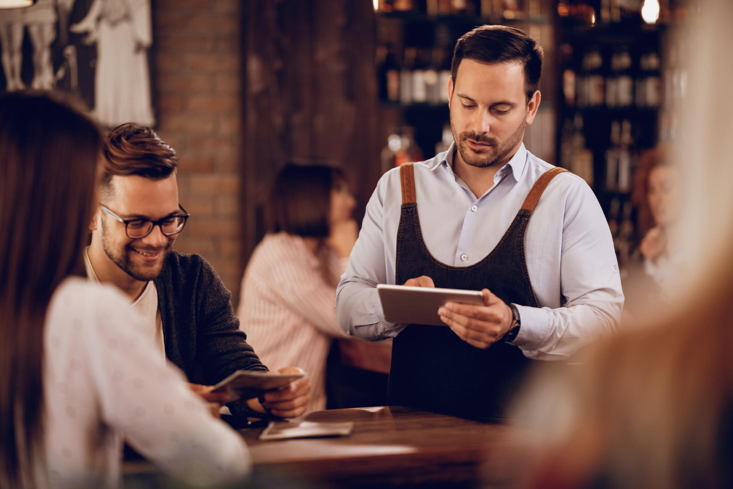 Serveur en tablier prenant une commande sur tablette auprès de clients souriants dans un restaurant chaleureux